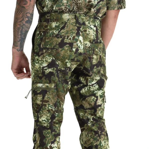 A person with a tattooed arm is shown from behind wearing Kryptek Valhalla Pants Obskura Transitional and shirt. The camouflage design includes shades of green, brown, and black. The person's arm is relaxed by their side.