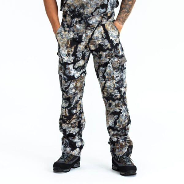 Person wearing Kryptek Stalker Pants Obskura Skyfall and black hiking boots. Their hands are in the pockets of the pants, and they have a tattoo on their visible left arm. The background is plain white.