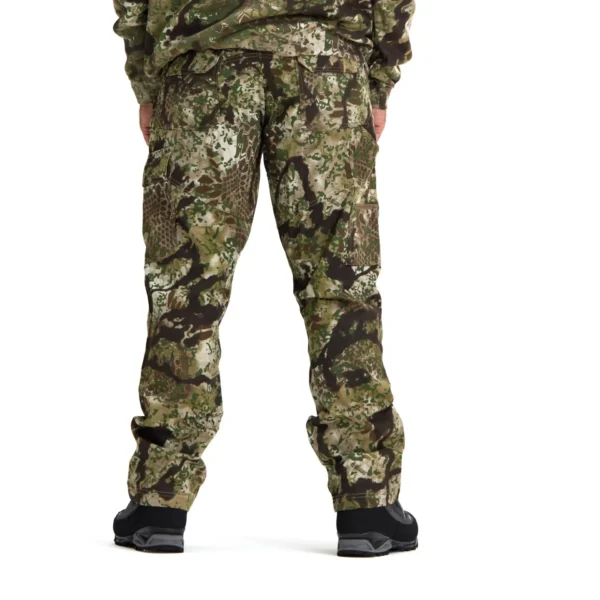 A person stands facing away, wearing Kryptek Stalker Pants Obskura Skyfall and a matching long-sleeved top. The outfit features various shades of green, brown, and beige. The person is also wearing black shoes or boots.