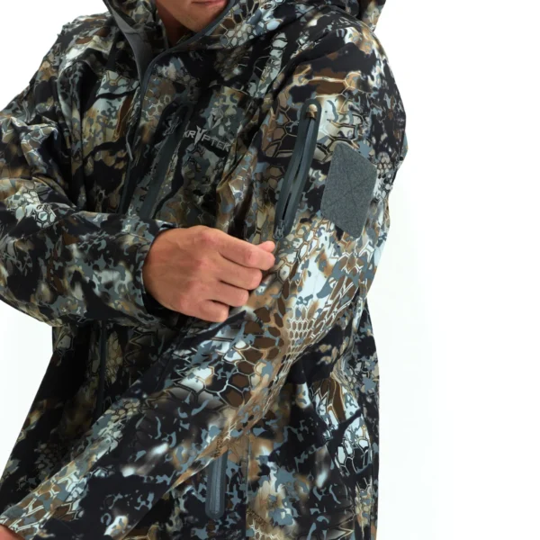 A person wearing a Kryptek Dalibor III Jacket Highlander adjusts the zipper on a sleeve pocket. The camouflage pattern is a mix of tan, green, and dark shades. The jacket also features a patch area on the sleeve. The background is plain white.