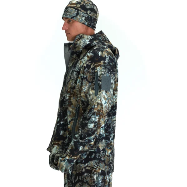 A person is standing sideways wearing a camouflage outfit, including a Kryptek Dalibor III Jacket Highlander, pants, gloves, and a beanie. The background is plain white, emphasizing the intricate pattern of the camouflage clothing.