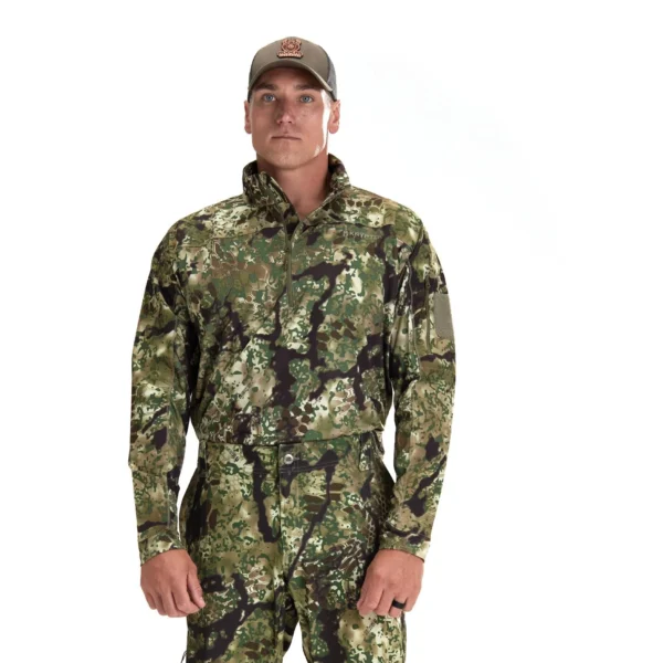 A man stands against a white background wearing a Kryptek Valhalla Long Sleeve Zip Highlander, including matching pants. He also wears a gray cap with a patch on the front. His arms are relaxed at his sides, and he looks straight at the camera.
