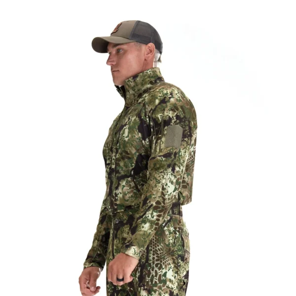 A person in profile view is dressed in a Kryptek Valhalla Long Sleeve Zip Highlander with long sleeves and a high collar. They are also wearing a black and grey baseball cap. The background is plain white.