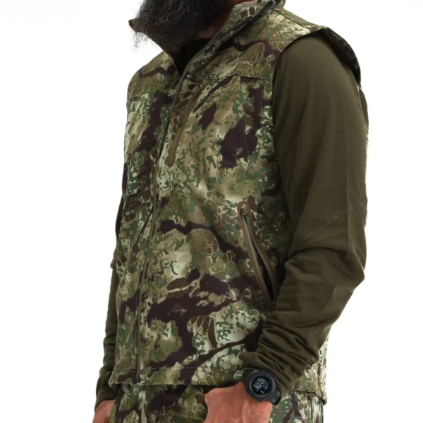 A person is wearing a Kryptek Dalibor Vest over a long-sleeve olive green shirt. They have a long beard and are also sporting a wristwatch with a black band on their left wrist. The image captures them from the waist up, oriented slightly to the right.