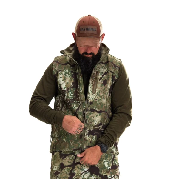 A man with a beard, wearing a Kryptek Dalibor Vest, olive green long-sleeved shirt, camouflage pants, and a brown cap, is adjusting his vest's zipper. He has a tattoo on his fingers and is standing against a plain white background.
