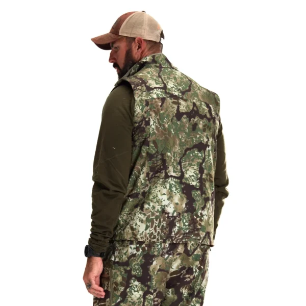 A man with a beard wears a khaki and green camouflage Kryptek Dalibor Vest and pants, and a brown and beige baseball cap. He stands with his back turned and head slightly to the side, giving a side profile view. He also wears a dark green long-sleeve shirt underneath the vest.