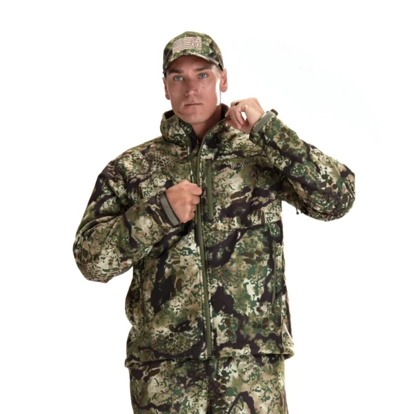 A person stands against a white background, wearing a Kryptek Njord Windproof Jacket, pants, and hat. The person is looking directly at the camera while adjusting the zipper of their jacket with their right hand.