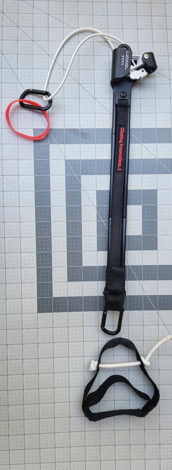 An Saka Foldable with a long black strap, a metal clip, and a coiled cable with a red and a black loop at each end, laid out on a gray grid-patterned surface.