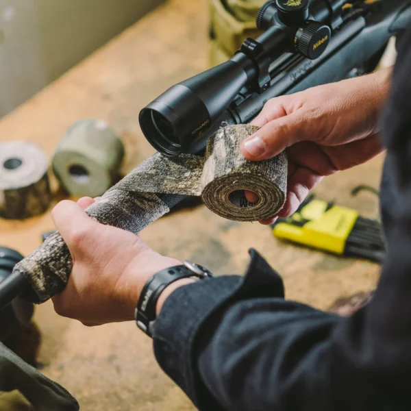 A person wrapping Camo Form Reusable Fabric Wrap around the barrel of a rifle on a workbench. Various hunting equipment, including rolls of Camo Form Reusable Fabric Wrap and tools, are visible in the background. The person is wearing a dark long-sleeve shirt and a watch on their left wrist.