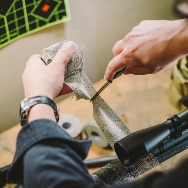 Two hands, wearing a dark jacket and a watch, use the Camo Form Reusable Fabric Wrap to cut a piece of camouflage fabric. A rifle scope and other materials are visible on the table. The workspace is seen from above, with a colorful target in the background.