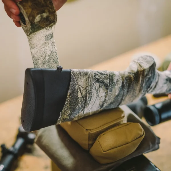 A person wraps a Camo Form Reusable Fabric Wrap around the stock of a rifle, which is resting on a beige shooting rest. Various tools and equipment are visible in the blurred background on a wooden surface.
