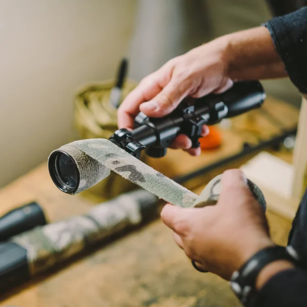 Two hands are shown wrapping Camo Form Reusable Fabric Wrap around a gun scope. The background is blurred, showing various tools and objects on a wooden workbench. The focus is on the hands and the scope being wrapped with tape.