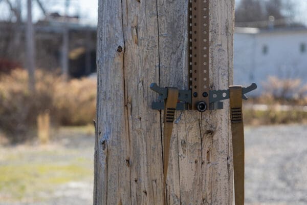 A close-up view of a weathered wooden utility pole with metal brackets and brown Ape Canyon Outfitters 3 Step Aider attached to it. The background is blurred, showing a mix of industrial and natural elements with grass and some buildings.