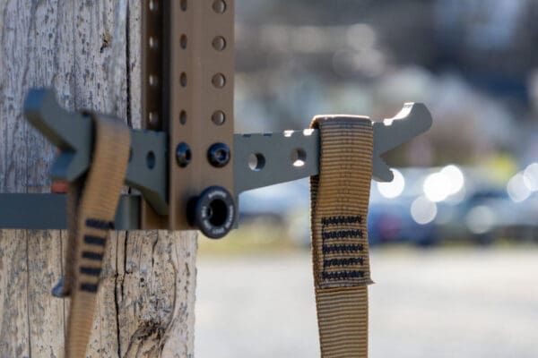 A close-up image of a mounted Ape Canyon Outfitters 3 Step Aider attached to a vertical wooden post with two brown straps secured to it. The background is blurred, showing an outdoor scene with soft lights and indistinct shapes of structures and greenery.