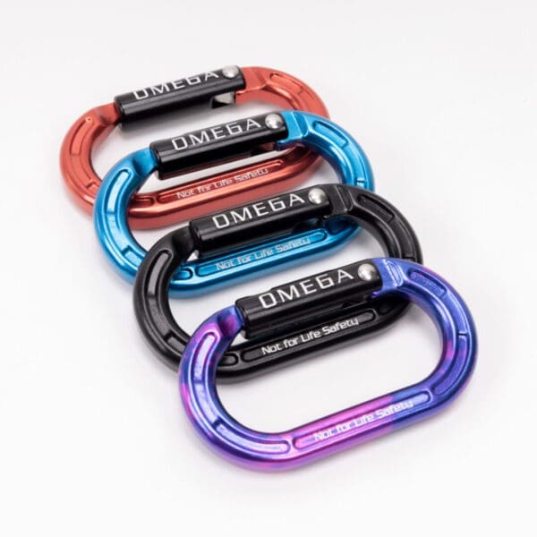 A display of five colorful carabiners arranged in a staggered row on a white background. The carabiners are labeled "Omega Pacific Micro Raider Aluminum Keylock Straightgate" and have a safety warning that reads "Not For Climbing". The colors from top to bottom are red, blue, black, teal, and purple.