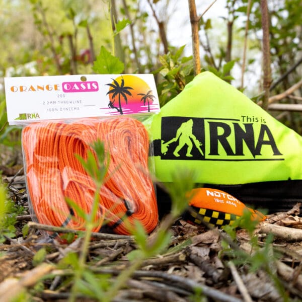 A package of Ape Canyon Outfitters 2.2mm Orange Oasis and a green-yellow throw weight with a black tree and climber logo, both branded "RNA," are set on the ground among green plants and twigs.