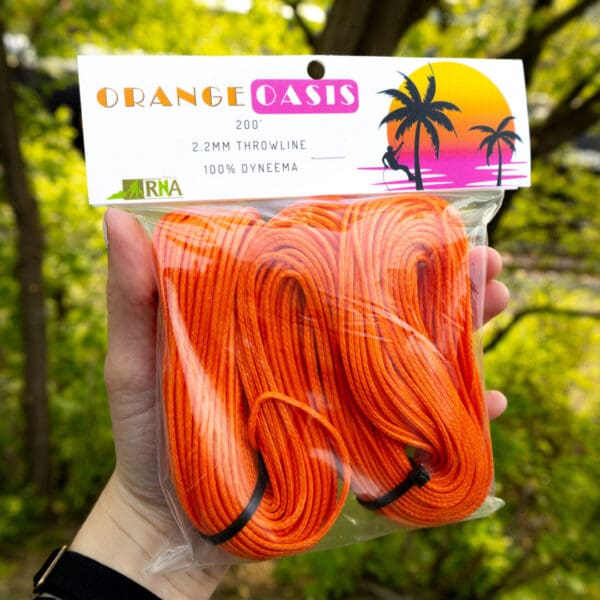 A hand holds a packet labeled "Ape Canyon Outfitters 2.2mm Orange Oasis" containing 200 feet of 2.2mm throwline made of 100% Dyneema. The throwline is bright orange. The packet's design features a sunset with palm trees and a person swinging from a tree.