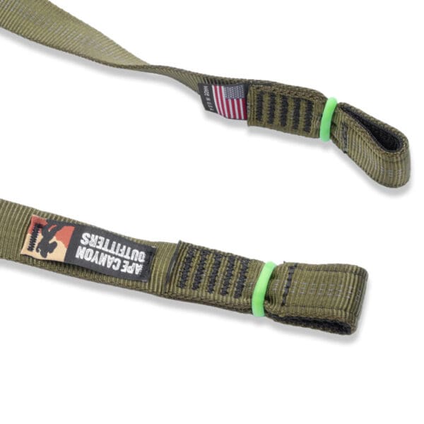 Close-up image of a green, heavy-duty strap with a black label showing a logo and the text "Ape Canyon Outfitters 3 Step Aider." There's also a small label with an American flag. The strap has reinforced stitching and a loop end secured with green rubber bands.