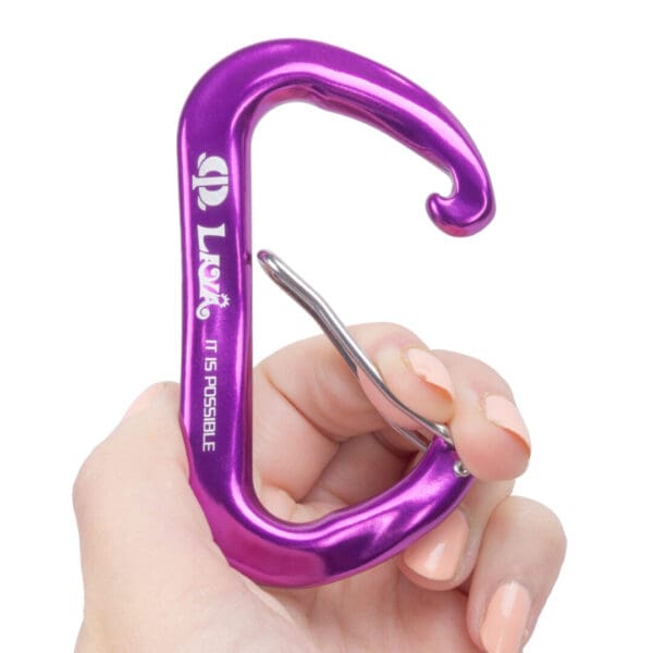A hand with light-colored nail polish holds a bright purple carabiner with a silver clasp. The carabiner has the text "Omega Pacific Lava Wiregate" and "IT IS POSSIBLE" printed on its side. The background is plain white.