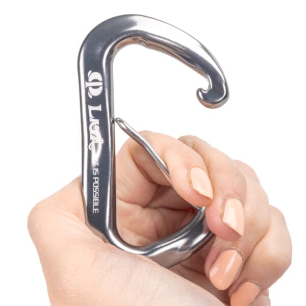 A hand with light skin tone holds an Omega Pacific Lava Wiregate, featuring the word "LOVE" and the phrase "IS POSSIBLE" engraved on the side. The carabiner has a unique, open shape with a smooth finish.