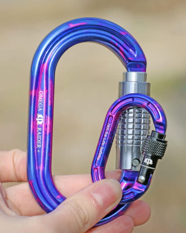 A close-up image of a shiny blue and purple Omega Pacific Micro Raider Aluminum Keylock Screw-Lok held in someone's hand. The carabiner has a silver, textured locking mechanism and intricate details visible, designed for secure connections in climbing and rigging.