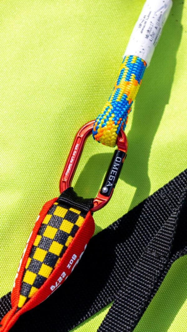 Close-up of a red climbing carabiner labeled "Omega Pacific Micro Raider Aluminum Keylock Straightgate" connecting a colorful rope with a yellow and blue pattern to a strap, against a bright green background. The carabiner is also attached to a woven black and red safety tether.