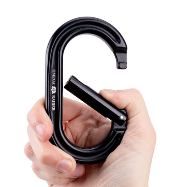 A close-up of a hand holding a black Omega Pacific Raider Aluminum Keylock Straight Gate - Black, with the Omega logo visible on its side. The carabiner is shaped like the letter "G" and appears to be made of metal. The thumb is pressing on the gate, which is partially open.