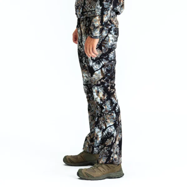Person wearing Kryptek Valhalla Pant Obskura Skyfall and hiking shoes, facing left. The pants feature a camouflage pattern with dark, light, and earthy tones, and the shoes are green with black accents. Only the lower half of the body is visible against a plain white background.