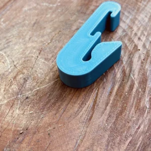 A blue, 3D-printed Paracord Saddle Aid lies on a worn, textured wooden surface. The Paracord Saddle Aid is shaped in an unconventional, angular design and has a smooth finish. The wood shows various scratches and lines, indicating frequent use or weathering.