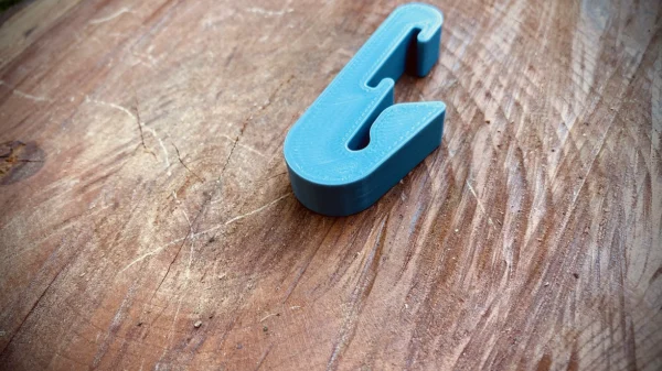 A blue, 3D-printed Paracord Saddle Aid lies on a worn, textured wooden surface. The Paracord Saddle Aid is shaped in an unconventional, angular design and has a smooth finish. The wood shows various scratches and lines, indicating frequent use or weathering.