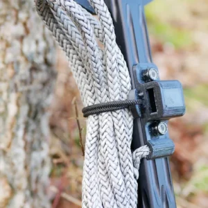 Close-up of a One Stick Clip & Hitch coiled and tied to a black metal bracket attached to a tree trunk. The tree's bark texture and some out-of-focus greenery in the background are visible. The One Stick Clip & Hitch appears to be used for securing or pulling purposes.