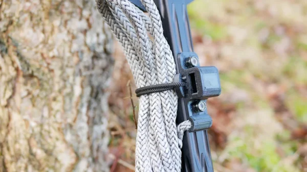 Close-up of a One Stick Clip & Hitch coiled and tied to a black metal bracket attached to a tree trunk. The tree's bark texture and some out-of-focus greenery in the background are visible. The One Stick Clip & Hitch appears to be used for securing or pulling purposes.
