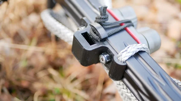 Close-up of a black and gray braided rope secured with a knot around the One Stick Clip & Hitch attached to a metal rod or bar. The background is blurred with earthy tones, likely suggesting an outdoor setting.