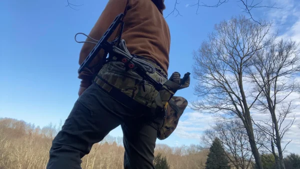 A person stands outdoors with their back turned, wearing a brown hoodie and gray pants. They have a camouflaged fanny pack with various gear, including a glass water bottle on their left side and One Stick Clip & Hitch on their right side, against a backdrop of a clear blue sky and bare trees.