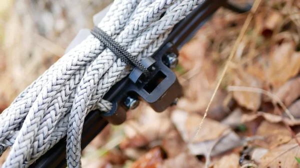Close-up of a bundle of thick gray braided rope secured with a One Stick Clip & Hitch and attached to a black metal bracket. The background consists of dry leaves and grass, indicating an outdoor setting.