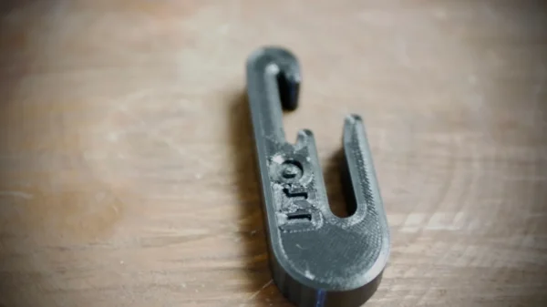 A close-up of a black, 3D-printed plastic tool with a hook and a U-shaped cutout. The Stick Hitch & Receiver Combo is placed on a wooden surface. The word "LIFT" is engraved on the top of the tool.