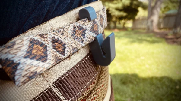 Close-up of a person wearing a patterned belt with a Paracord Saddle Aid attached to it. In the background, there is a grassy area with trees and fencing, suggesting an outdoor, backyard setting.