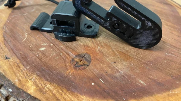 Two One Stick Clip & Hitch are shown on a wooden surface. One is lying flat, while the other is resting partially on the first clasp, showcasing their design and functionality. The background is a natural, textured wooden surface with visible grain patterns.