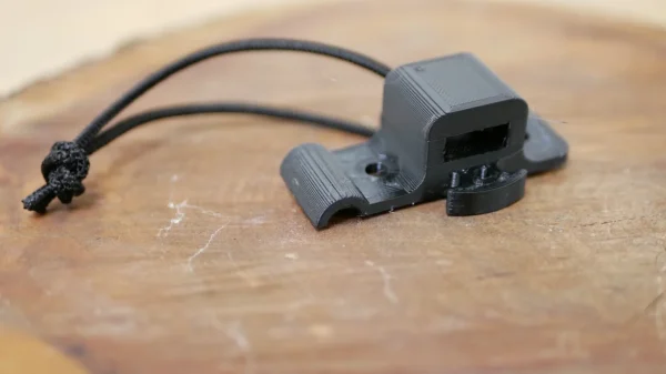 A black 3D-printed object lies on a wooden surface. It appears to be a custom-designed One Stick Clip & Hitch with a small looped string attached to one end and an opening or slot on the other. The object likely serves a specific functional purpose.