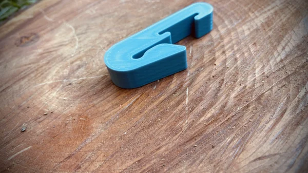 A small, blue 3D-printed Paracord Saddle Aid lies on a textured wooden surface. The surface has visible grain patterns and small scratches.