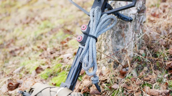 A close-up of climbing equipment placed against a tree stump. The gear includes the One Stick Clip & Hitch with two red knobs and a coil of gray rope. The ground is covered with brown leaves and patches of green moss. A part of a khaki-colored bag is visible at the bottom.