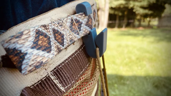 Close-up of a woven outdoor chair in a grassy backyard. The chair features a Paracord Saddle Aid secured with a blue plastic clip, and intricate stitching details on its body. Trees and a fence are visible in the blurred background.