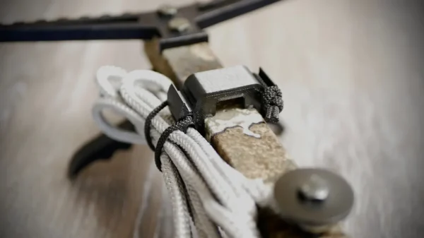 Close-up shot of a Stick Hitch & Receiver Combo, showing paracords neatly secured around the handle, a small saw-like blade, and a screw. The tool rests on a light wooden surface.