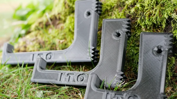 Three black 3D-printed plastic brackets with ridged edges, imprinted with the letters "ITO," are leaned against a moss-covered tree trunk. The Pocket Dewclaw Hanger (3 Hangers, Amsteel, and Locker) are positioned on grass, showcasing their design and detailing.
