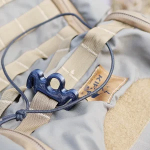 A close-up of a gray backpack with tan straps, featuring a blue cord lock threaded through a black string on the front. The fabric appears sturdy, suitable for outdoor activities. The background is slightly blurred, suggesting a natural setting. This is The Molle BOO (Bungee Operated Object).