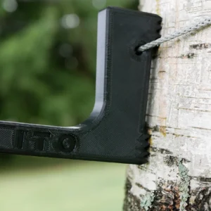 A black plastic Pocket Dewclaw Hanger (3 Hangers, Amsteel, and Locker) hooks onto a birch tree, holding a gray rope. The hanger has the letters "ITO" imprinted on it. The background is a blurred green landscape with soft natural light.