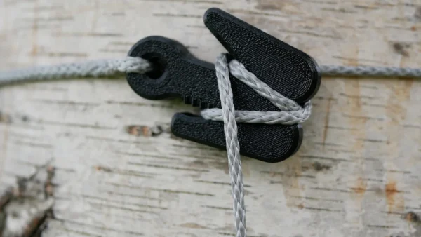 Close-up of a gray rope tied around The Pocket Dewclaw Hanger (3 Hangers, Amsteel, and Locker) attached to a light-colored, textured wooden surface. The clip helps secure the rope tightly in place. The background showcases the rough texture and natural patterns of the wood.