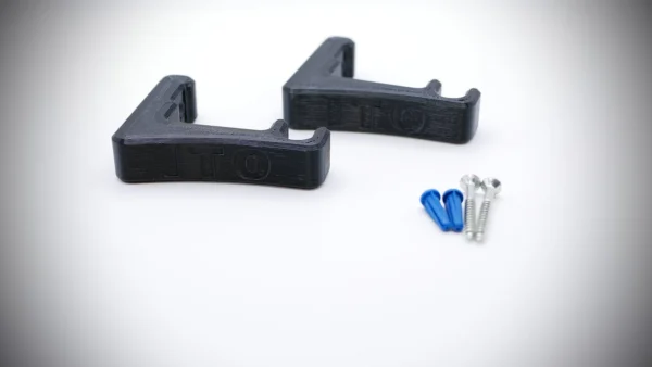 Two black, L-shaped 3D-printed Stick Bow Wall Hangers are placed on a white surface beside two blue wall anchors and two screws. The brackets have the letters "ITO" embossed on them. The items are arranged neatly and the image has a soft gradient background.