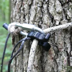 Close-up of a tree trunk with several ropes tied around it using a Flexi Ohm Tether Locker and a black plastic clip. The ropes are securing something out of the frame in a wooded area. The background shows a dense forest.