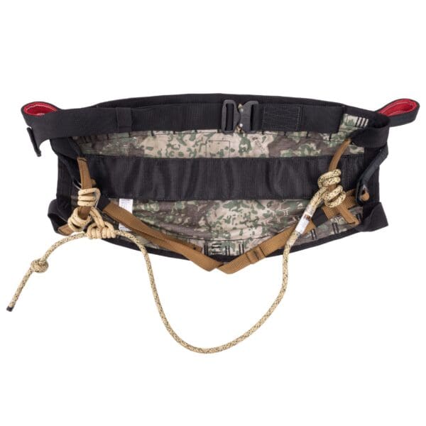 The Ape Canyon Outfitters Wood Deluxe Saddle Obskura Transitional is a rock climbing waist harness with black padded straps, a green and beige camouflage-patterned interior, red loops, and multiple black buckles. It features thick beige ropes tied in knots and loops for additional safety and support.